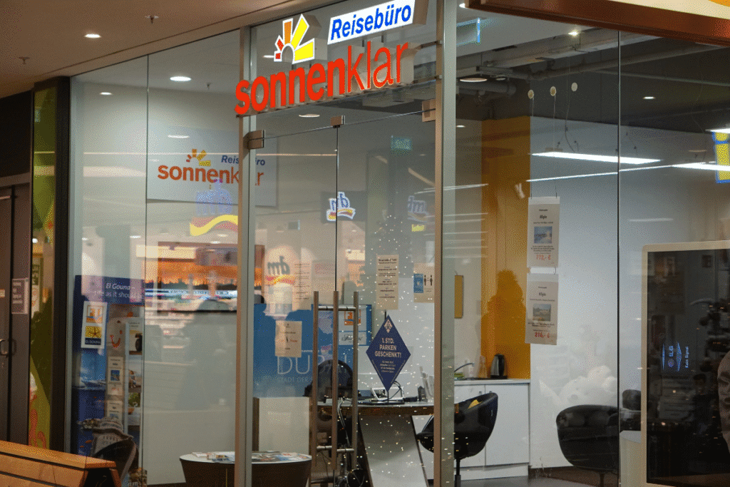 Glasfront eines Reisebüros mit rotem Schriftzug 'sonnenklar' und blauem Schild 'Reisebüro' darüber, Innenraum mit Stühlen und Tischen sichtbar.